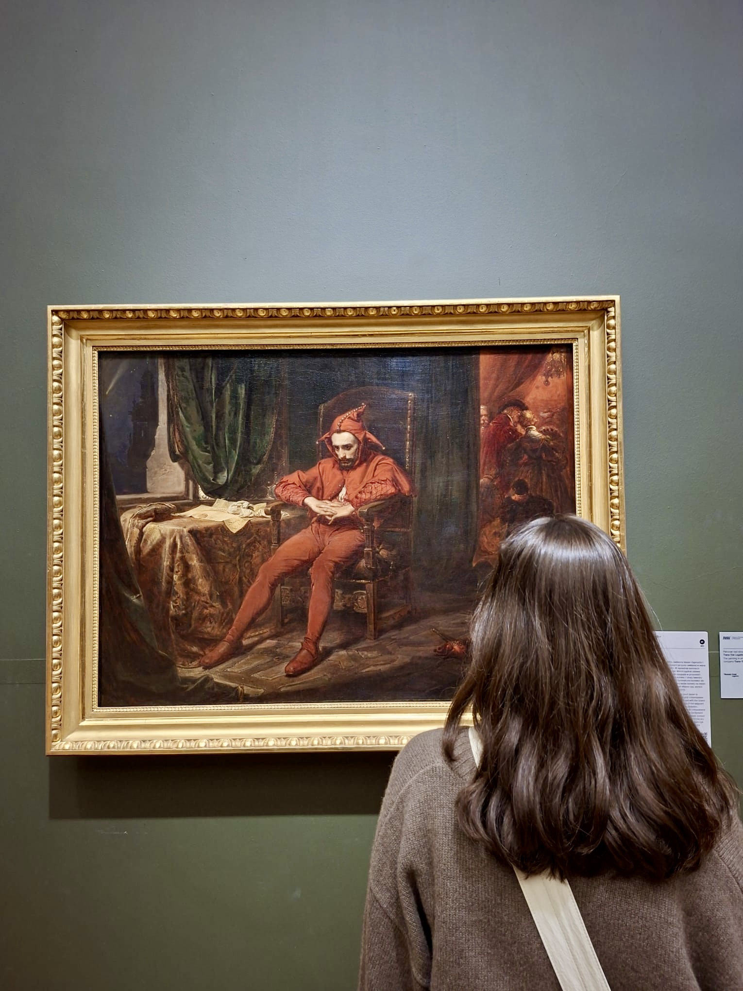 A visitor seen from behind viewing a framed 19th-century painting of a seated jester dressed in red, hung on a deep green gallery wall.