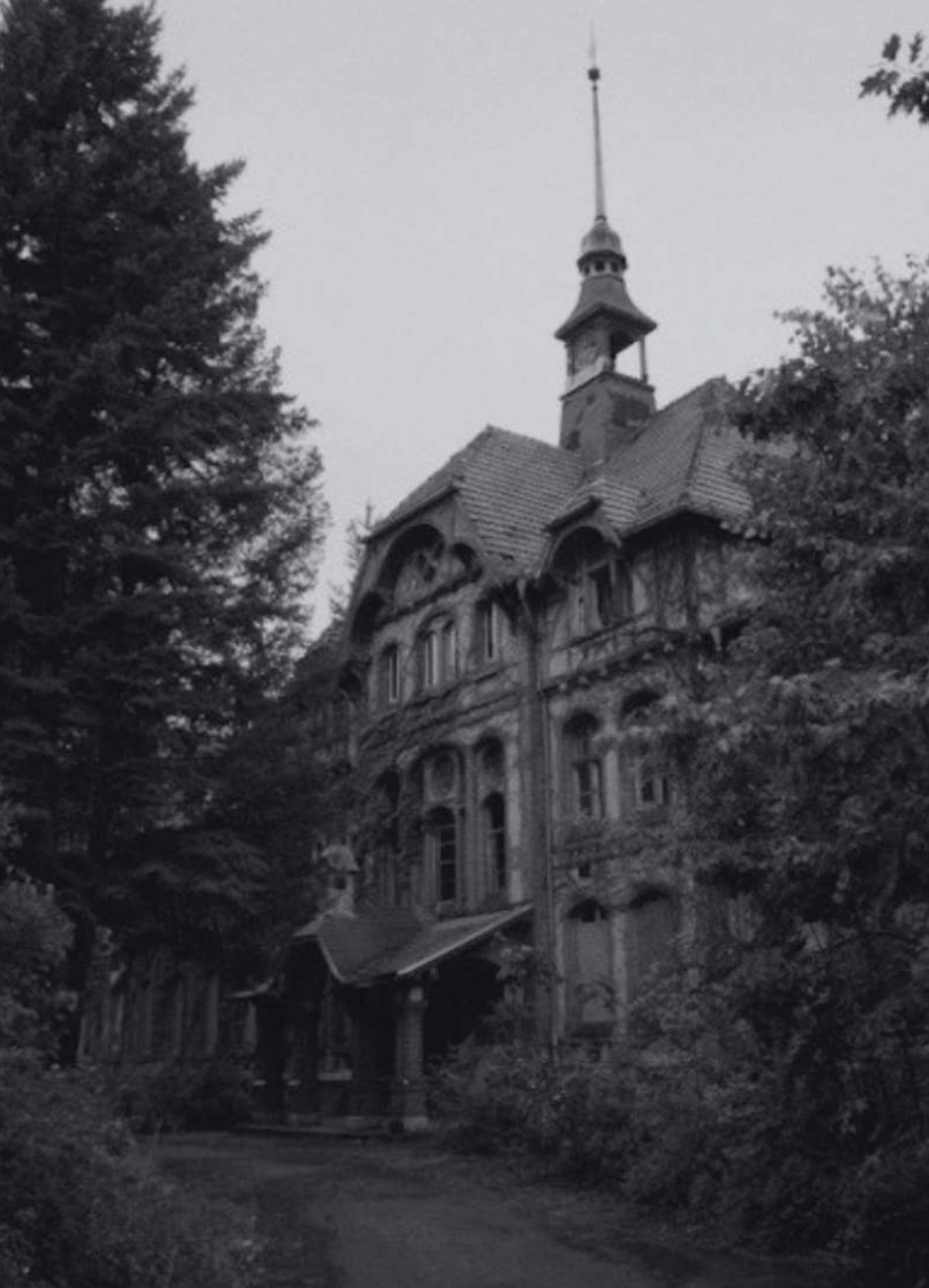 Black-and-white photograph of the abandoned Whitney Houston Haus at Beelitz-Heilstätten: an overgrown, decaying historic sanatorium building with a tall spire, framed by trees.