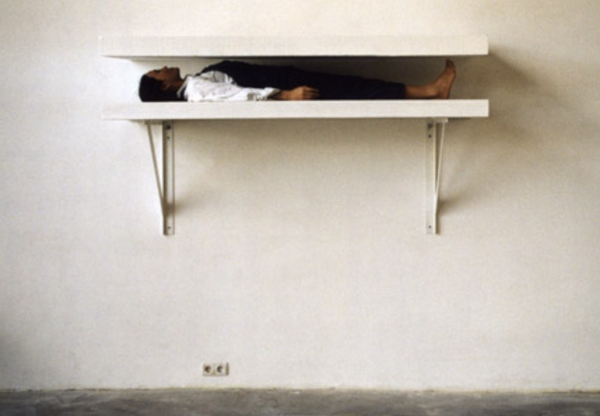 Performance still: the artist Yingmei Duan lies fully enclosed between two white wall-mounted shelves.