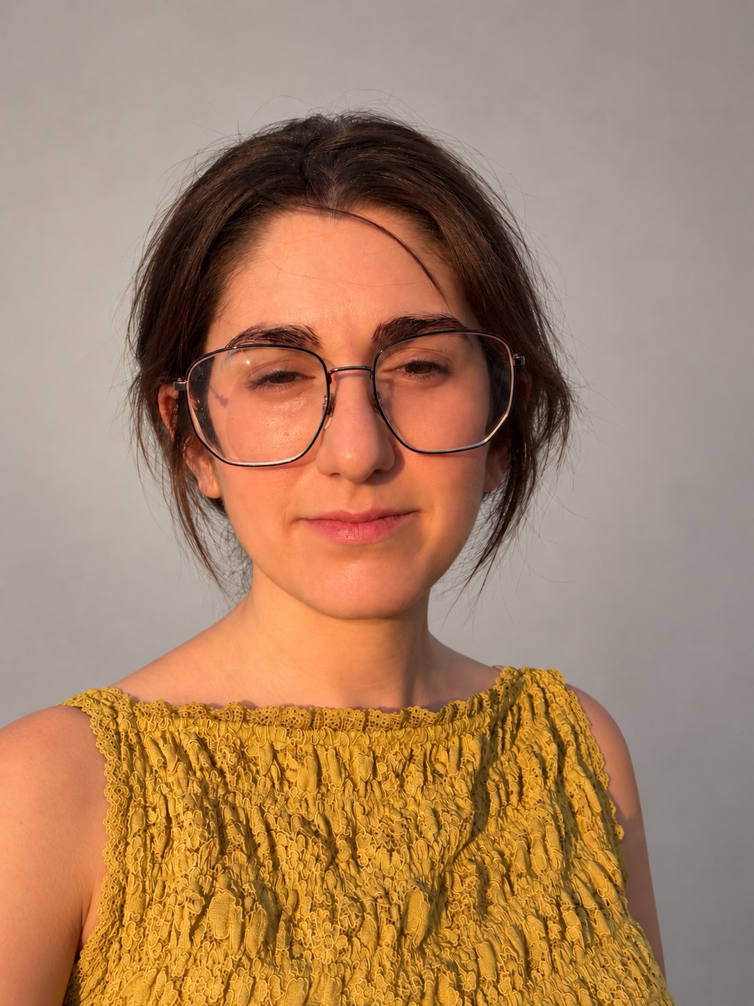 Portrait of Madeline Heller wearing large wire-framed glasses and a mustard-yellow textured sleeveless top, against a pale grey wall in warm light.