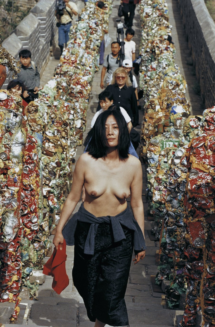 Performance documentation: the artist walks bare-chested up a long flight of steps lined with columns of crushed, multicoloured cans and debris, with other people following behind.