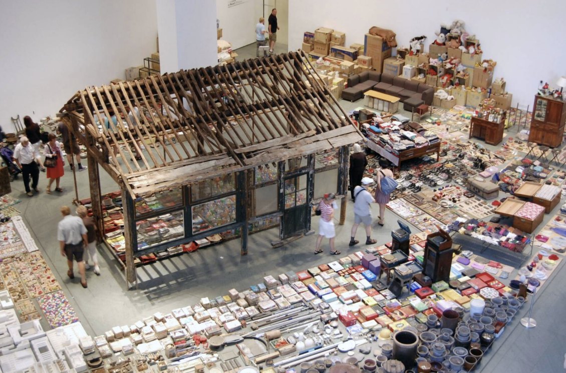 Gallery installation: a weathered wooden house frame surrounded by thousands of everyday household objects (clothing, shoes, bottles, boxes, and utensils) laid out in neat rows across the floor as visitors walk among them.