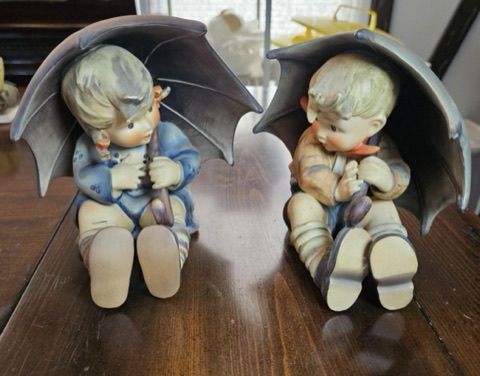 The Umbrella Girl and Umbrella Boy Hummel figurines sitting side by side on a wooden table, each sheltering under a large grey umbrella.