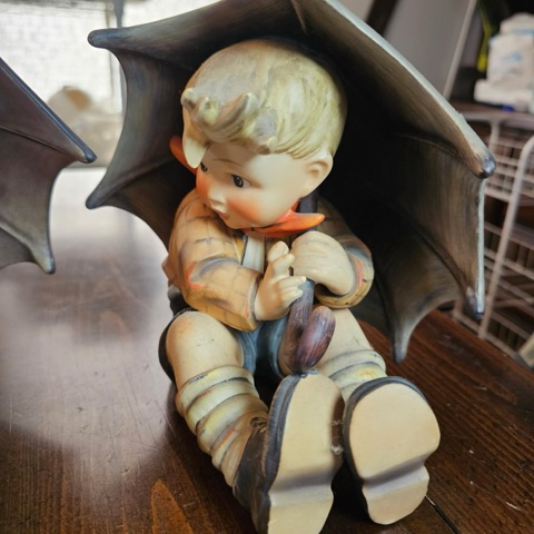 Close-up of the Umbrella Boy figurine in a striped jacket and orange scarf, clutching his umbrella handle, with striped socks and worn boots.