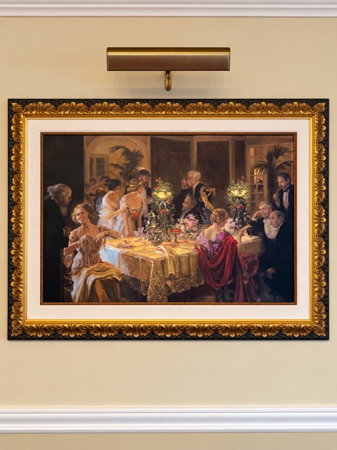A large painting of an elegant candlelit dinner party in an ornate gold frame with a cream mat, hung beneath a brass picture light.