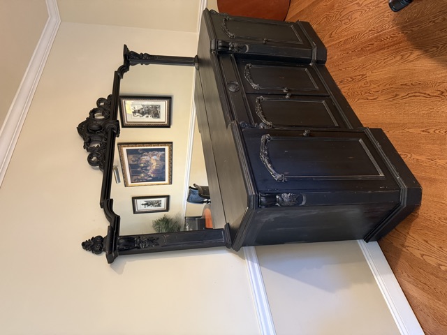 A black distressed sideboard with carved arched doors and a tall carved mirror on top, reflecting framed art across the room.