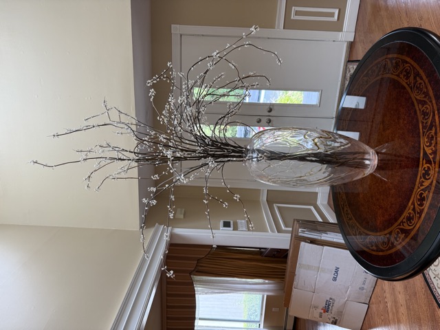 A tall clear glass vase holding white blossom branches on a round inlaid wooden table in a bright entry hall.