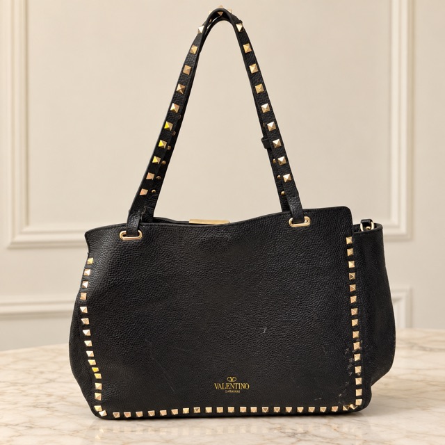A black leather Valentino Garavani tote bag edged with gold pyramid studs on a marble surface.