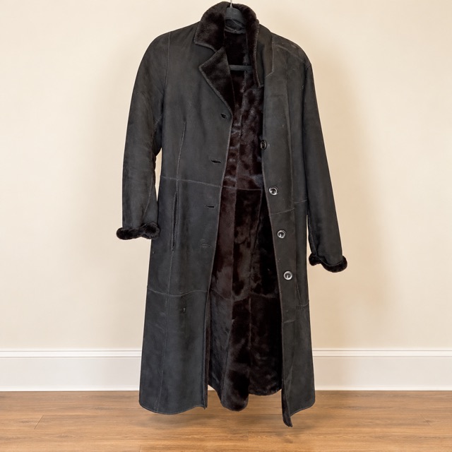 A long dark brown shearling coat with a plush fur lining, hanging against a cream wall.