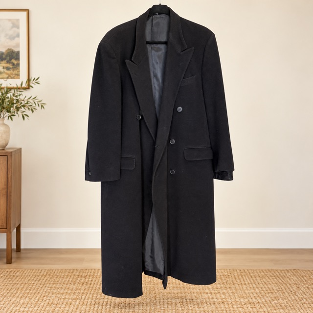 A long black double-breasted wool overcoat with peaked lapels, hanging in a softly lit room.
